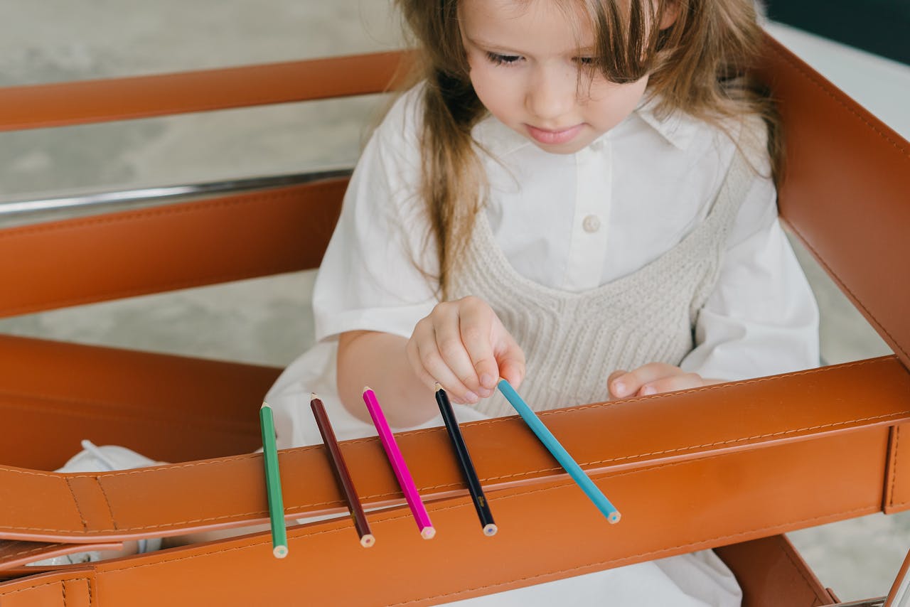 A young girl attentively arranging colored pencils on a leather chair.